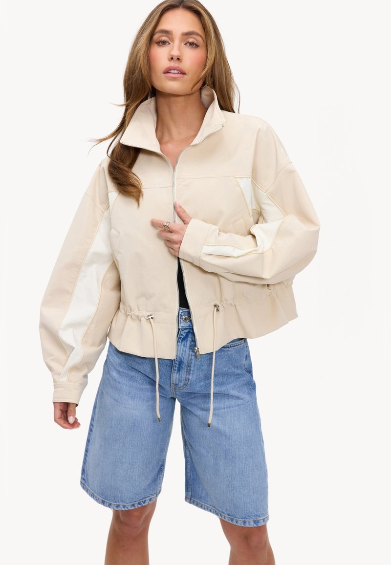 Femme portant une veste beige zippée avec cordons et un short en denim bleu clair jusqu'au genou sur fond blanc.