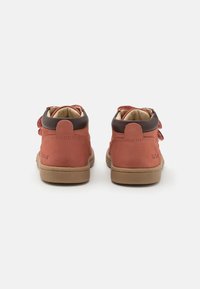 Stivaletti alla caviglia in suede rosso con dettagli in pelle marrone e punta tonda. Presentano una chiusura con cinturino in Velcro e una suola in gomma beige.