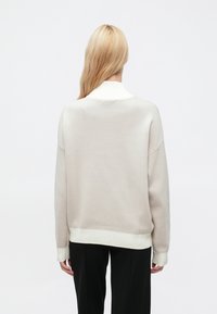 Világosbézs pulóver bordázott fehér gallérral és mandzsettával, laza szabású, eresztett vállú, fekete nadrággal párosítva.