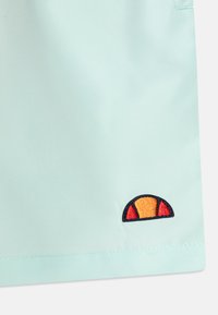 Svijetlo zeleni materijal sadrži izvezeni logo u crvenoj, narančastoj i crnoj boji, smješten u donjem kutu. Glatka tekstura, ležeran dizajn.