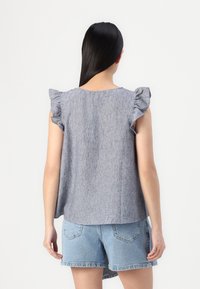 Blå chambray ermeløs topp med volanger på skuldrene, myk tekstur og avslappet passform, paret med lyse denimshorts.