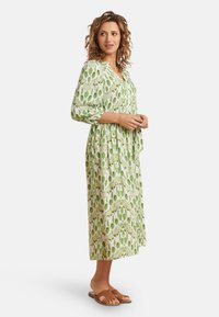Kleid mit grünem Blätter-Muster, dreiviertel Ärmeln, V-Ausschnitt und gerafftem Bund. Getragen mit braunen flachen Sandalen.