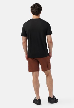 Schwarzes T-Shirt mit kurzen Ärmeln, kombiniert mit braunen Shorts, die über eine seitliche Tasche verfügen, eine glatte Textur haben und slim fit sind. Schwarze Sportschuhe runden das Outfit ab.
