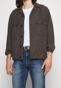 Giacca button-up grigio scuro con due tasche sul petto, colletto aperto e maniche arrotolate, abbinata a una t-shirt bianca e jeans blu.