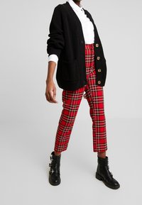 Černý pletený cardigan s dvěma kapsami a knoflíkovým zapínáním, zkombinovaný s červenými kostkovanými kalhotami a černými kotníkovými botami se stříbrným kováním.