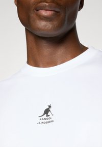 Hvid T-shirt med korte ærmer lavet af blødt stof, med et sort grafisk logo af en kænguru med en golfkølle og tekst nedenfor.