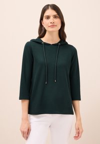 Dunkelgrüner, gerippter Hoodie mit einer Kängurutasche, Zugbändern mit Metallenden und Dreiviertelärmel, kombiniert mit weißen Hosen.