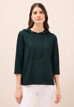Sudadera de color verde oscuro, con canalé, bolsillo canguro, cordones con extremos de metal y mangas tres cuartos, combinada con pantalones blancos.