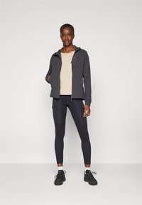 Schwarze Zip-Jacke, beiges Hemd und dunkle Leggings. Features ein sportliches Design, strukturierten Stoff und kontrastierende Panels. Schwarze Sneakers.