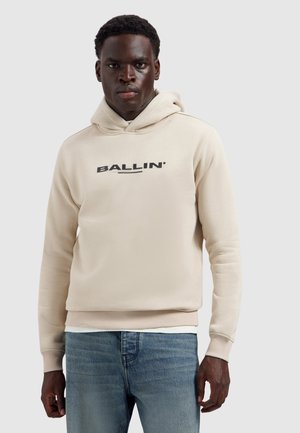 Man draagt een beige hoodie met de tekst "BALLIN", gecombineerd met een blauwe spijkerbroek, staand tegen een effen lichtgrijze achtergrond.