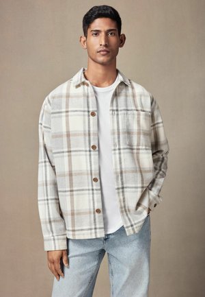 Plaid shirt in beige and gray tones, featuring a button-down front, chest pocket, and long sleeves, worn over a white t-shirt with light denim jeans.