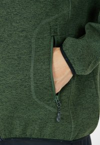 Giacca in pile verde con tasca laterale con zip, tessuto testurizzato e polsini neri a contrasto. Ideale per attività all'aperto.