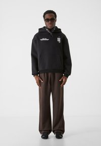 Sort hættetrøje med oversized pasform, hvid tekstdetalje, frontlomme, kombineret med brune vide bukser og sorte sneakers. Glat stoftekstur.
