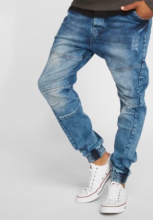 Man met lichtblauwe joggerbroek van spijkerstof met elastische boorden en witte lage sneakers, staand tegen een effen achtergrond.