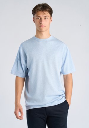Jonge man die recht vooruit kijkt, gekleed in een lichtblauw T-shirt met korte mouwen en een donkere broek, staande tegen een effen lichte achtergrond.