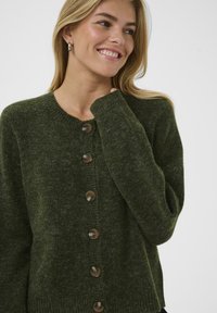 Zaļš adīts kardigāns ar apaļu kakla izgriezumu, pieciem brūniem pogām priekšpusē, ribotām piedurknēm un mīksta teksturēta auduma.