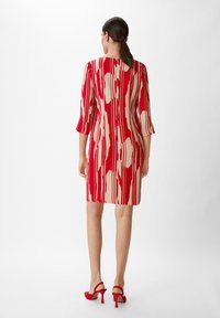 Rotes und beiges gemustertes Kleid mit geradem Schnitt, Dreiviertelärmeln und knielanger Länge. Getragen mit roten High Heels.
