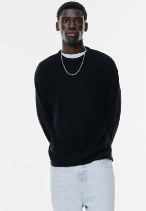 Black ribbed sweater with a crew neck, worn over a white shirt, paired with light blue denim jeans. Features a silver chain necklace.