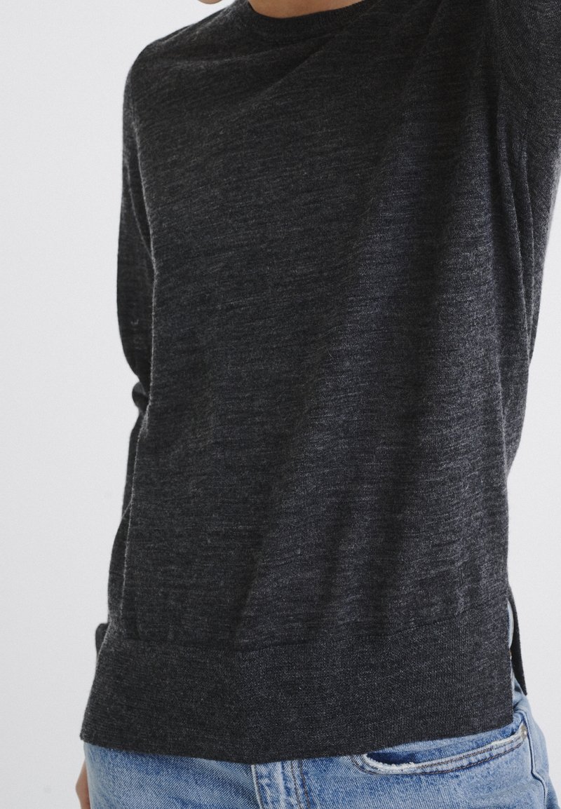 Pull gris foncé en tissu doux, avec un col rond, des manches longues et un ourlet côtelé. Porté avec un jean bleu clair pour créer un contraste.