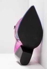 Botte en cuir rose à bout pointu avec semelle noire et talon bloc, positionnée à la verticale contre une surface d'angle blanche.