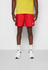 Rde kratke Nike z gladko teksturo, v kombinaciji z belimi nogavicami do gležnjev in športnimi čevlji z mornarskimi poudarki. Model nosi rumeno majico.