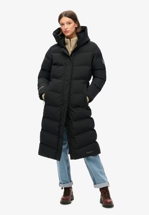 Femme debout, les mains dans les poches, portant un long manteau matelassé noir, un jean bleu clair retroussé, un pull beige et des bottines marron à lacets.