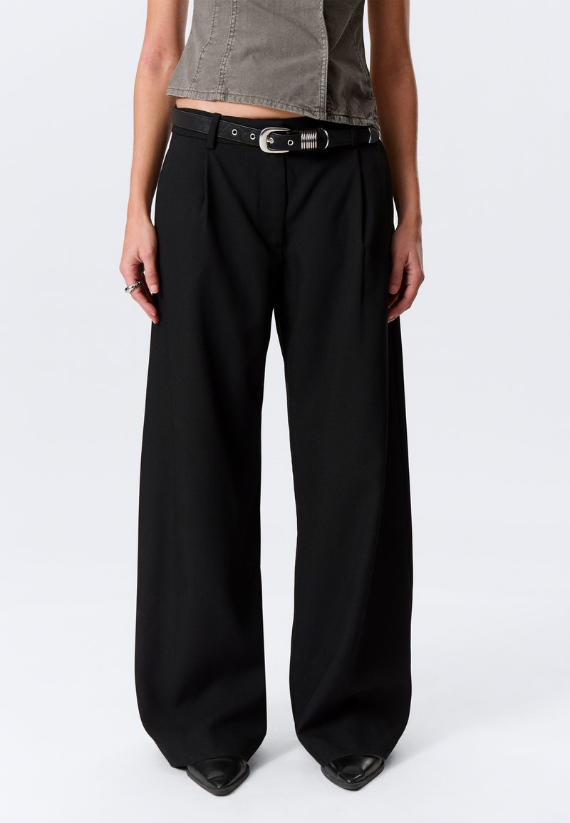 Sieviete tērpusies melnās platām bikšu kājām ar melnu jostu, dūmakaini brūnu īsu topu un melnām norādītām kurpēm uz vienkārša baltā fona.