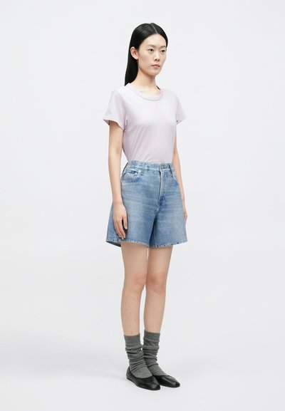 Mladá žena stojaca, oblečená v svetlofialovej krátkosleevej košeli, modrých rifľových šortkách, sivých ponožkách a čiernych plochých topánkach, na bielom pozadí.