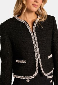 Czarna marynarka z tkaniny tweedowej o teksturowanym wykończeniu, z perłowymi akcentami przy kołnierzu i kieszeniach oraz dopasowaną sylwetką.