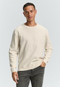Beiger Strickpullover mit langen Ärmeln, rundem Ausschnitt, gerippten Bündchen und lockerer Passform, kombiniert mit grauen Jeans. Weicher Stoff mit dezenten Nähten.