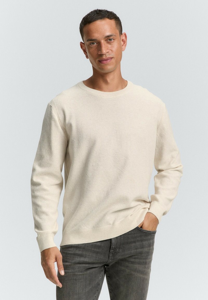 Beiger Strickpullover mit langen Ärmeln, rundem Ausschnitt, gerippten Bündchen und lockerer Passform, kombiniert mit grauen Jeans. Weicher Stoff mit dezenten Nähten.