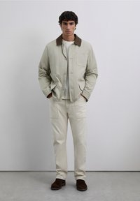 Joven de pie con las manos en los bolsillos, vistiendo una chaqueta beige con cuello marrón, pantalones color crema y zapatos marrones oscuros, frente a una pared lisa.