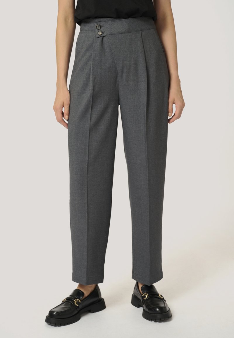 Femme portant un pantalon gris cintré taille haute avec deux boutons et des mocassins noirs avec boucles dorées, debout devant un fond uni.
