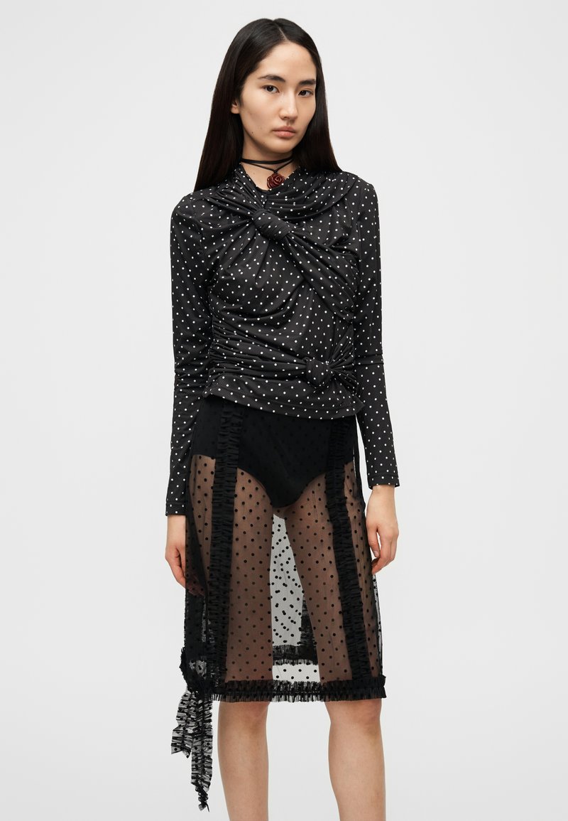 Haut noir à pois avec un détail noué, associé à une jupe noire transparente à pois, avec des bords froncés et une longueur mi-mollet.