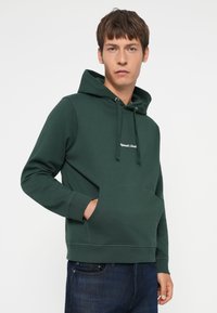 Haut à capuche en coton vert avec une poche avant, doté d'une capuche avec cordon de serrage, de poignets côtelés et d'un texte brodé sur la poitrine. Porté avec un jean foncé.