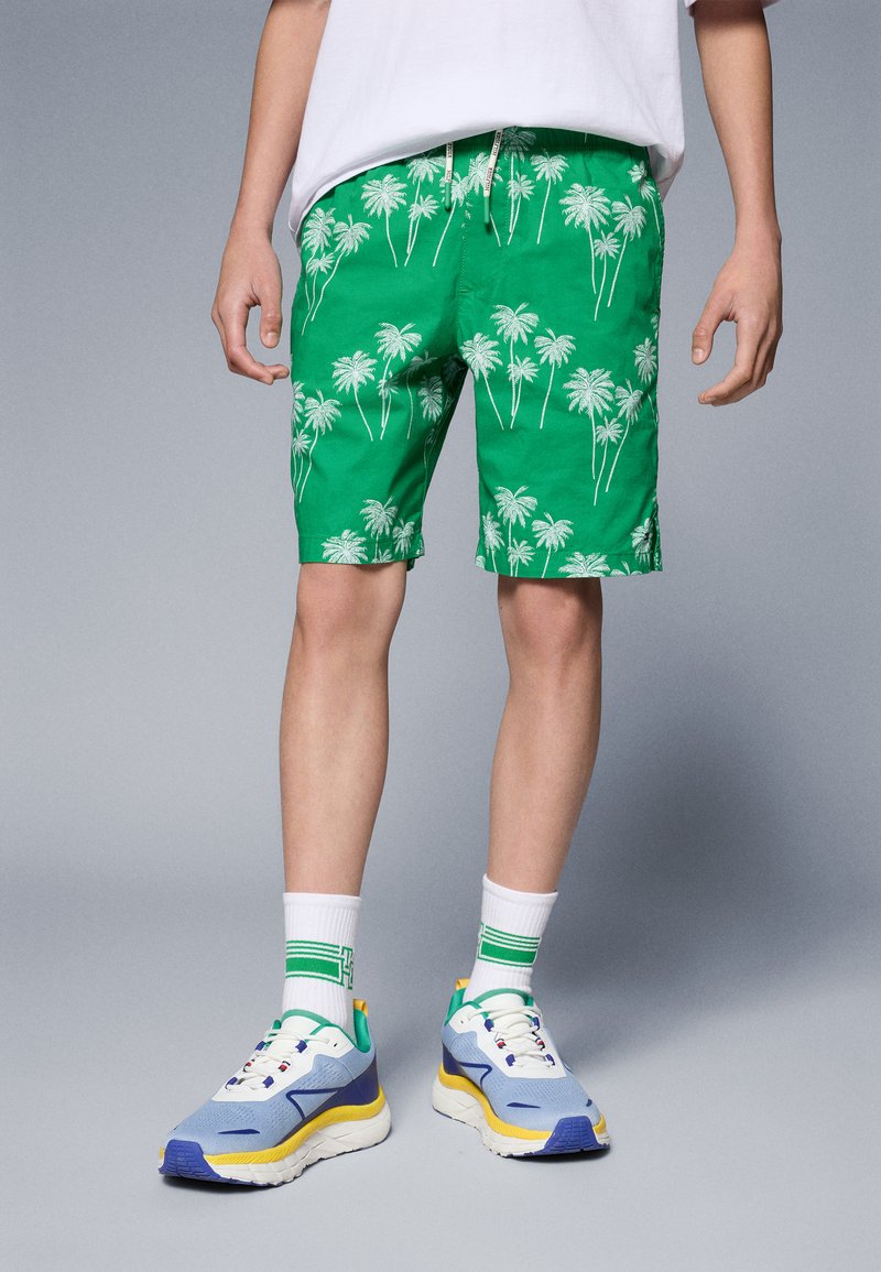 Gröna shorts med vita palmträdsmönster, lättviktsmaterial och resårmidja. Kombineras med blå, gula och vita sneakers.