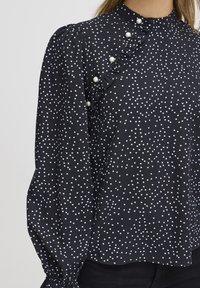 Blusa nera con pois bianchi, collo alto, dettaglio arricciato su un lato, accenti di perle e maniche lunghe a sbuffo con polsini elasticizzati.