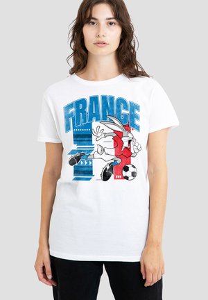 Junge Frau trägt ein weißes T-Shirt mit blauem und rotem France-Schriftzug und einem Cartoon-Hasen, der mit einem schwarz-weißen Ball Fußball spielt.