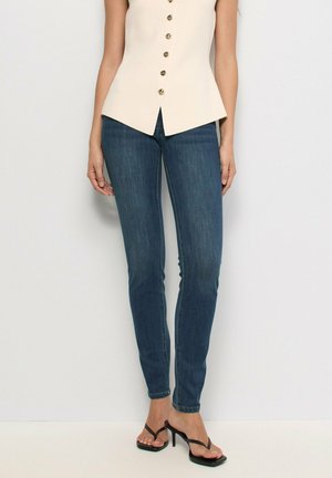 Vaqueros azul ajustados combinados con una blusa sin mangas beige con botones y sandalias negras de tacón sobre un fondo claro.