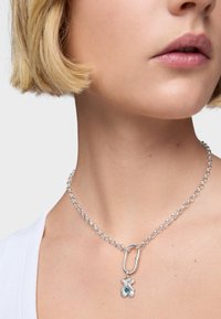 Srebrny naszyjnik łańcuszkowy z dużym ogniwem i zawieszką z niebieskim kamieniem szlachetnym, spoczywający na gładkiej szyi. Prosty wzór i wypolerowana tekstura.