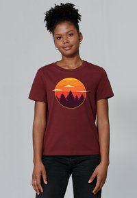 T-shirt en coton bordeaux avec un graphique de coucher de soleil circulaire aux teintes orange, jaune et violet, et des silhouettes d'arbres en bas.