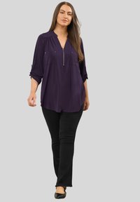 Langärmlige, tiefviolette Bluse mit Reißverschluss am Hals, zwei Brusttaschen und hochgerollten Ärmeln, kombiniert mit schwarzen, ausgestellten Jeans und Ballerinas.