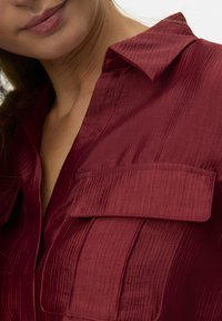 Bordeauxfarbiges Hemd aus strukturiertem Stoff mit leichtem Glanz, ausgestattet mit einem spitzen Kragen und zwei Brusttaschen.