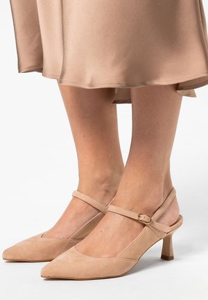 Talons aiguilles en daim beige avec des bouts pointus et de fines brides de cheville, associés à une jupe beige fluide se terminant au-dessus des genoux.