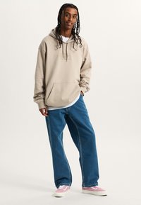 Felpa con cappuccio beige, dotata di tasca frontale e patch con logo, abbinata a jeans larghi blu e sneakers rosa, su uno sfondo neutro.