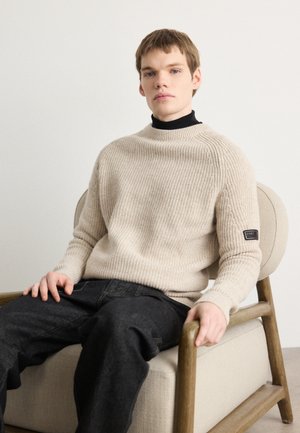Beige geripptes Strickoberteil mit hohem Kragen und Logo-Patch, kombiniert mit dunklen Jeans, getragen während sie in einem Holzstuhl sitzt.