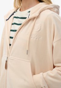 Hoodie zippé pêche clair avec une poche avant, des cordons de serrage et un logo brodé. Porté par-dessus une chemise blanche à rayures vertes.