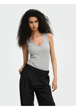 Jeune femme aux longs cheveux bouclés portant un haut gris sans manches à col en V et un pantalon noir taille haute, debout avec une main dans la poche.