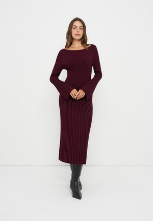 Femme portant une longue robe côtelée rouge foncé avec des manches évasées et des bottes noires à talons hauts, debout sur un fond blanc.