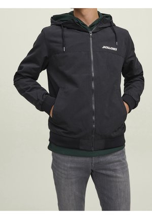Homme portant une veste noire zippée à capuche Jack & Jones par-dessus un sweat à capuche vert foncé et un jean gris, les mains dans les poches de la veste.